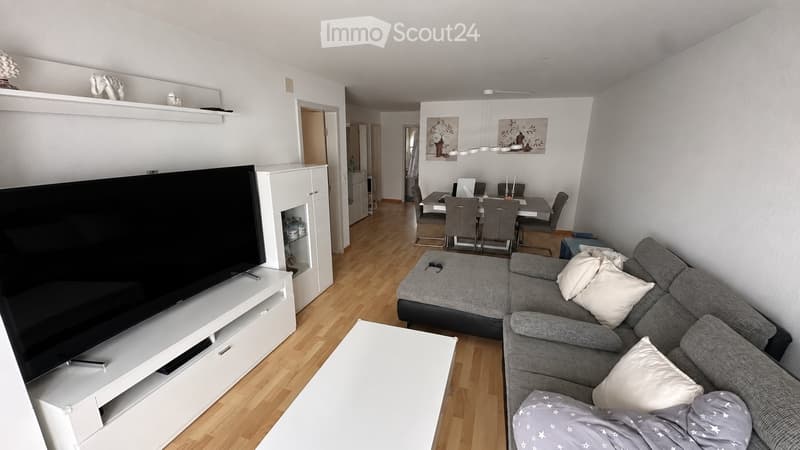 Modern living room with grey sectional sofa, white throw pillows, coffee table, flat-screen TV on white stand, dining area with table and chairs, and decorative shelves.