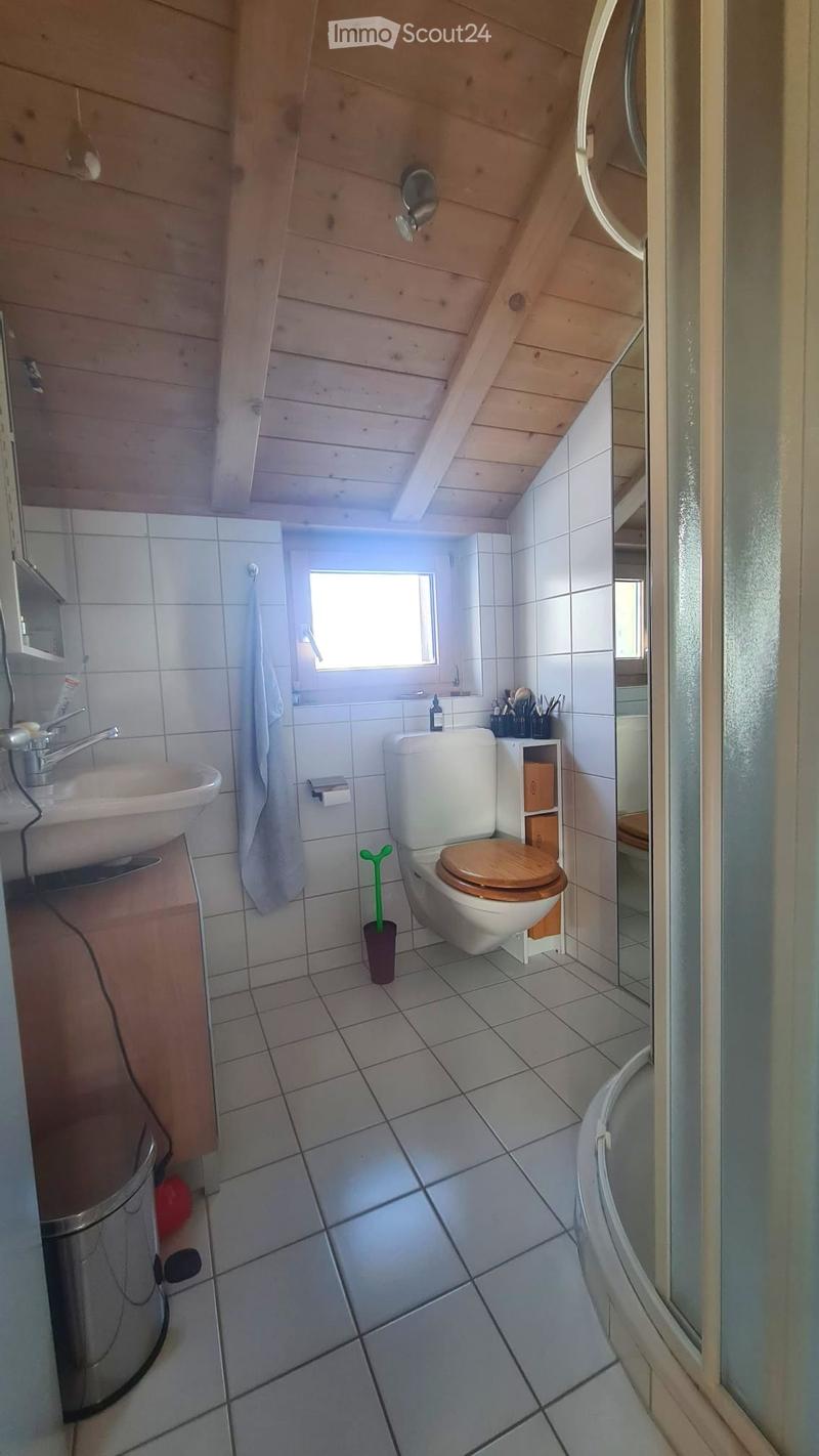 Bathroom with white tiled walls and floor, wooden ceiling beams, a sink with a faucet, a toilet with a wooden seat, a toilet brush, a mirror, a towel, and a small cupboard with toiletries.