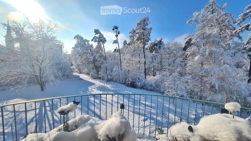 A snowy outdoor balcony overlooking a snow-covered forest and a distant church spire, with a metal railing and snow-covered furniture.