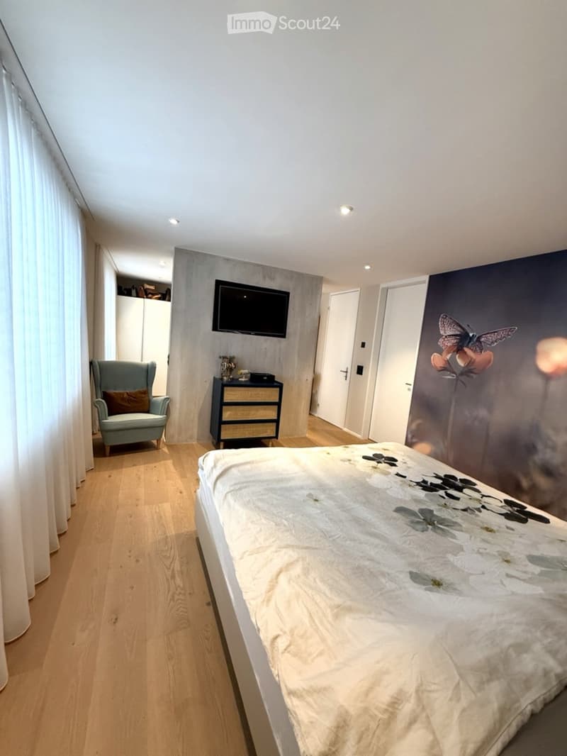 Modern bedroom with a large bed, a light blue armchair, a nightstand, and a wall-mounted TV. The room features wooden flooring and large windows with white curtains.
