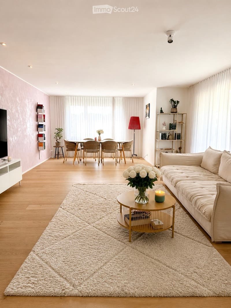 Modern living room with light wooden flooring, white walls, and large windows covered by sheer curtains. Features a cream-colored sofa, a round coffee table with a vase of white flowers, a dining area with a table and chairs, a floor lamp, and a shelf wit