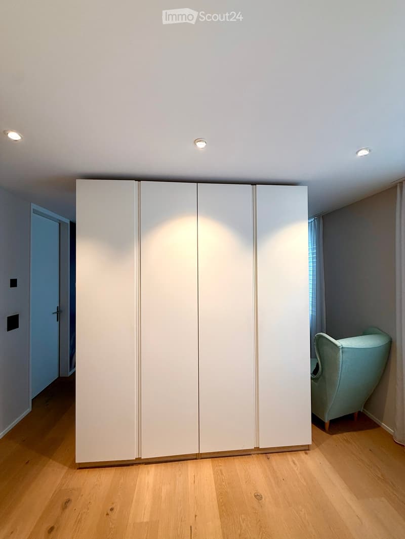 A modern, minimalist room with a large white sliding wardrobe and a light blue armchair near a window with curtains.