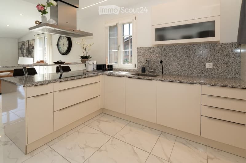 Modern kitchen with white cabinets, granite countertops, and a central island. Features include a sink, faucet, coffee maker, clock, vase with flowers, and chairs.