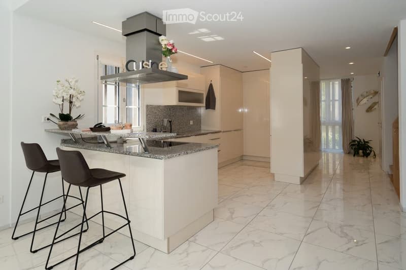 Modern kitchen with white cabinets, granite countertops, and a central island with two high stools. Features a stainless steel range hood, a sink, and decorative vases on the counter.