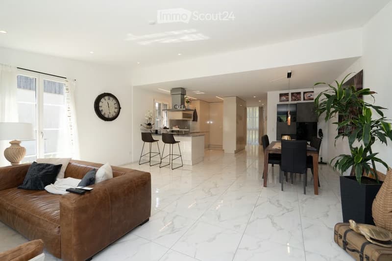 Open-plan living and dining area with a brown leather sofa, a dining table with chairs, and a kitchen area with bar stools. The space features white walls, marble flooring, and large windows with sheer curtains.