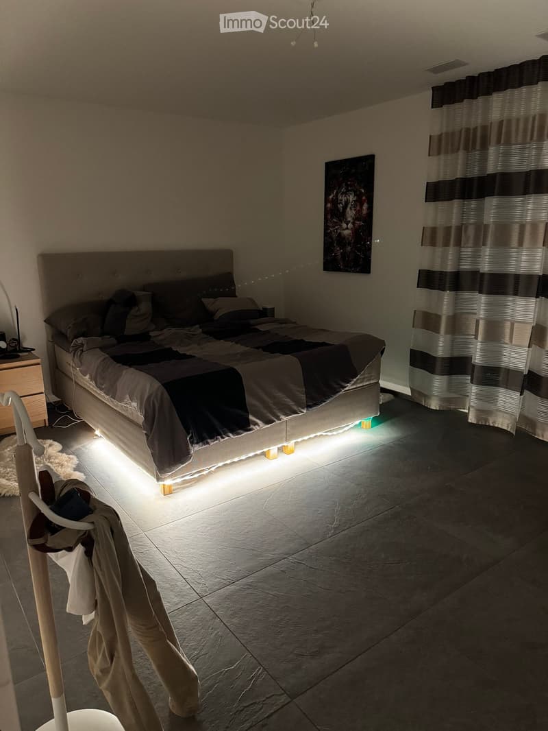 Modern bedroom with a large bed, nightstand, painting, and a curtained window. The room features tiled flooring and ambient lighting under the bed.