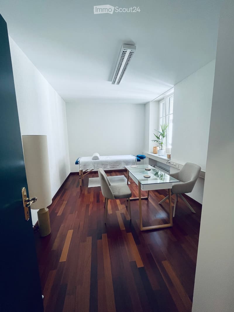 Bright, minimalist room with wooden flooring, a glass table, two chairs, a floor lamp, a small plant, and a treatment table with a white sheet.