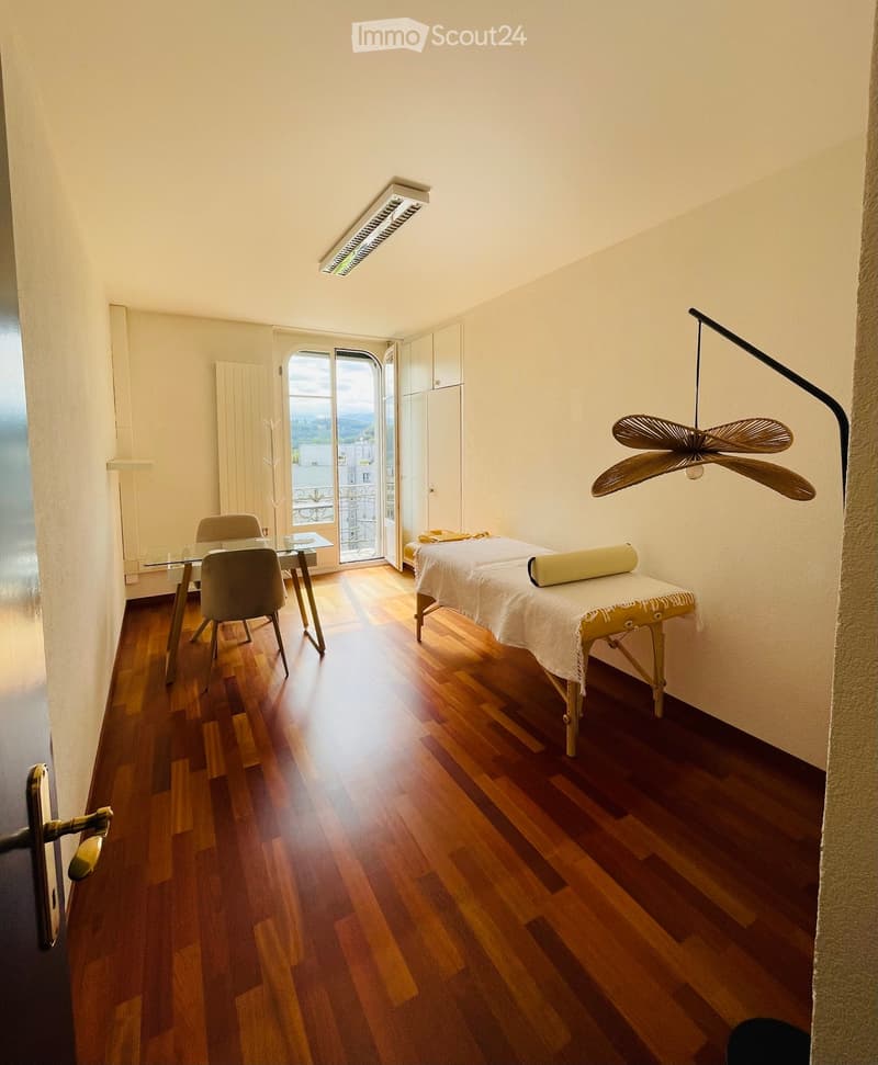 A well-lit room with wooden flooring, a massage table, a glass table with chairs, and a hanging fan.