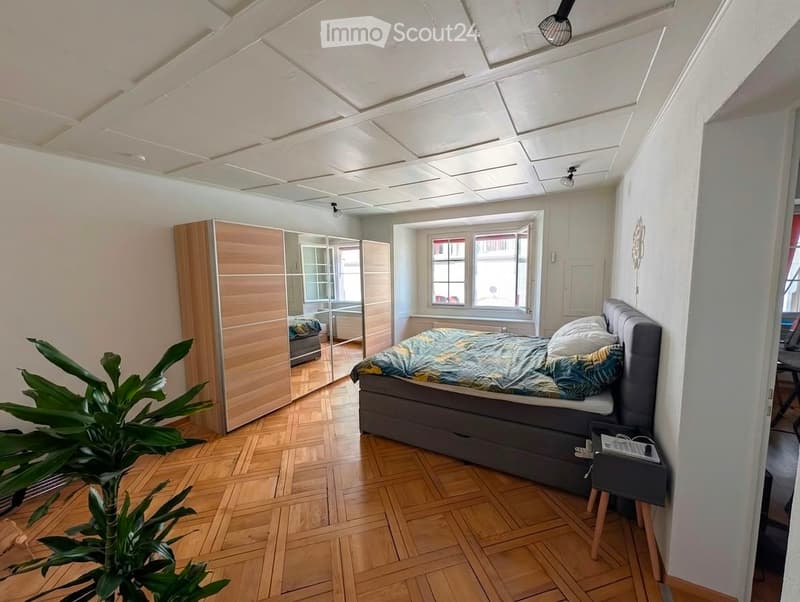 Modern bedroom with wooden flooring, large window, grey upholstered bed, nightstand, wardrobe, and a leafy plant in the corner.