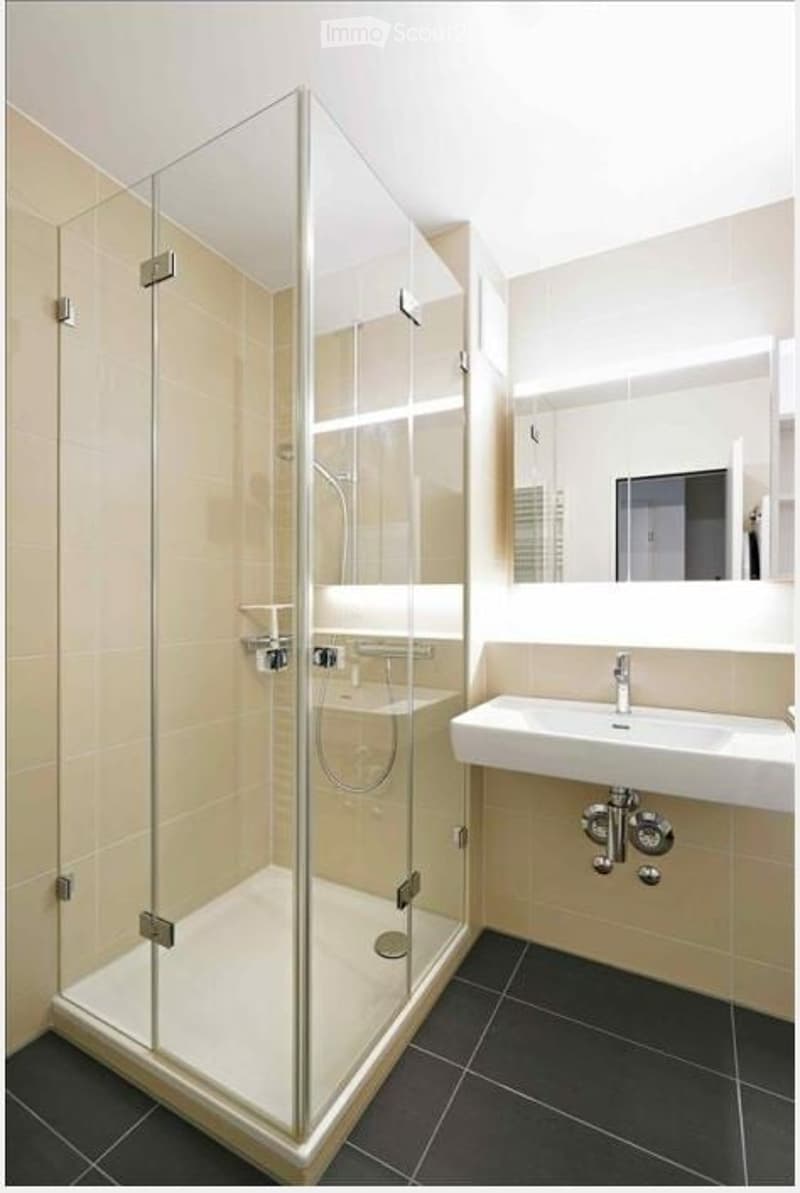 Modern bathroom with a glass-enclosed shower, a white sink, and a large mirror. The walls are tiled in a light beige color, and the floor is dark tiled.
