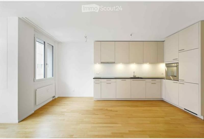 Modern kitchen with light-colored cabinets, a sink, and an oven. The floor is made of light wood, and the walls are white.