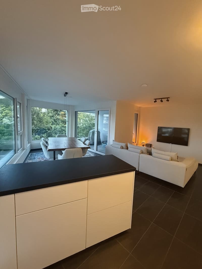 Open-plan living and dining area with a white sofa, coffee table, dining table with chairs, and a kitchen island. Large windows offer a view of greenery outside.