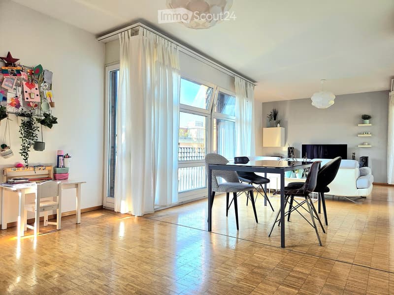 Bright living and dining area with wooden flooring, large windows, and modern furniture including a sofa, dining table, chairs, desk, and shelves.