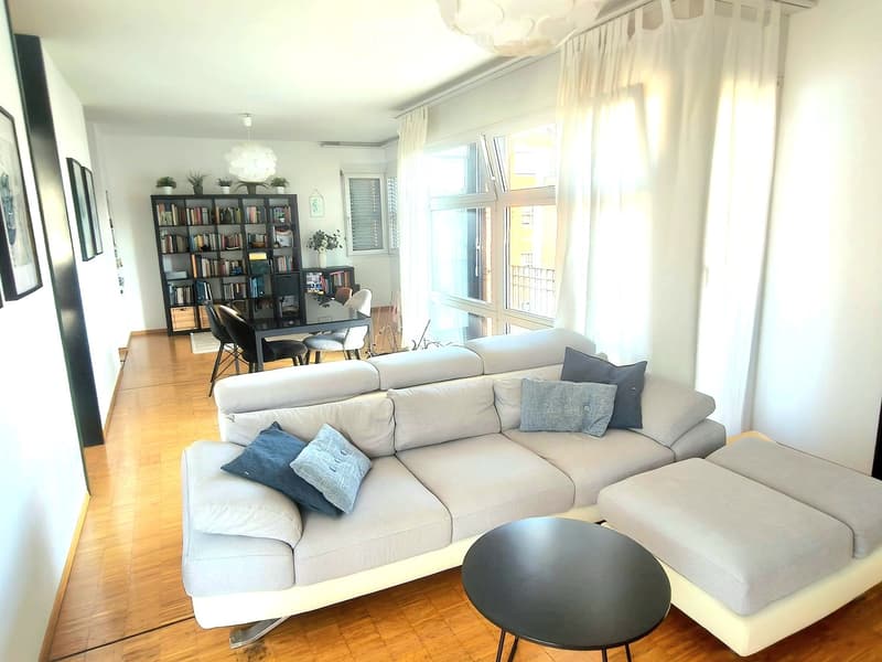 Modern living room with a grey sectional sofa, coffee table, dining area with chairs and table, bookshelf, and large windows with white curtains.