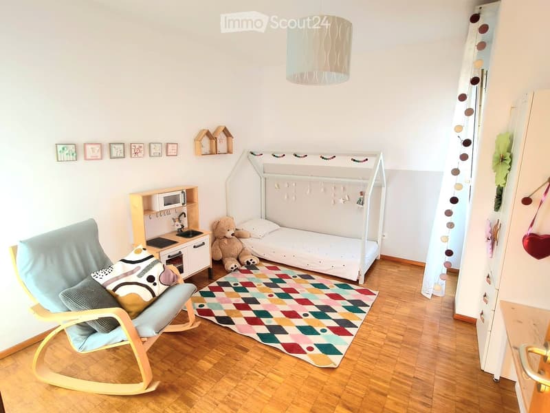 A cozy children's bedroom with a white loft bed, a rocking chair, a small table with a microwave, and a colorful rug.