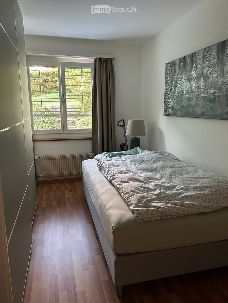 Bright bedroom with wooden flooring, a neatly made bed, a bedside lamp, and a large window with curtains offering a view of greenery.