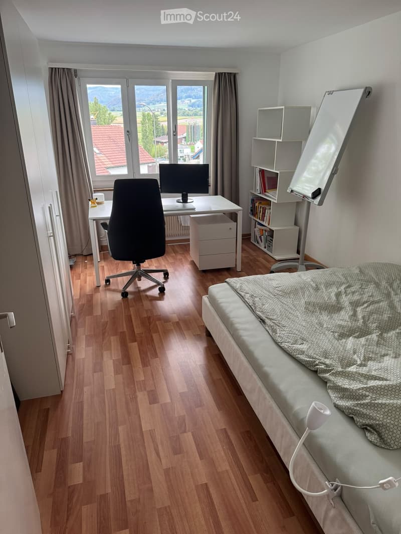 Well-lit bedroom with a bed, desk, chair, and bookshelf. Large windows offer a scenic view.