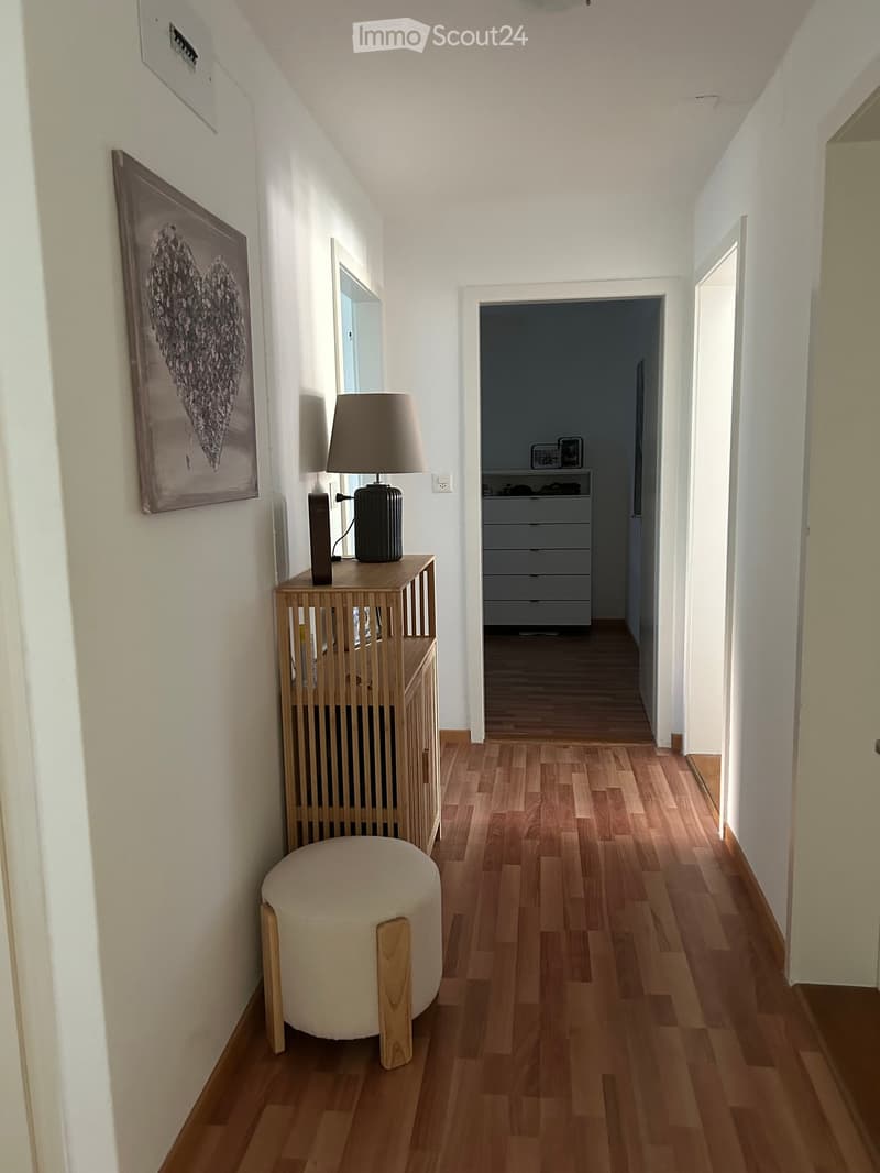A narrow hallway with wooden flooring, white walls, and a small wooden table with a lamp and picture frame on it. A round stool is placed near the table, and a doorway leads to another room with a dresser visible.