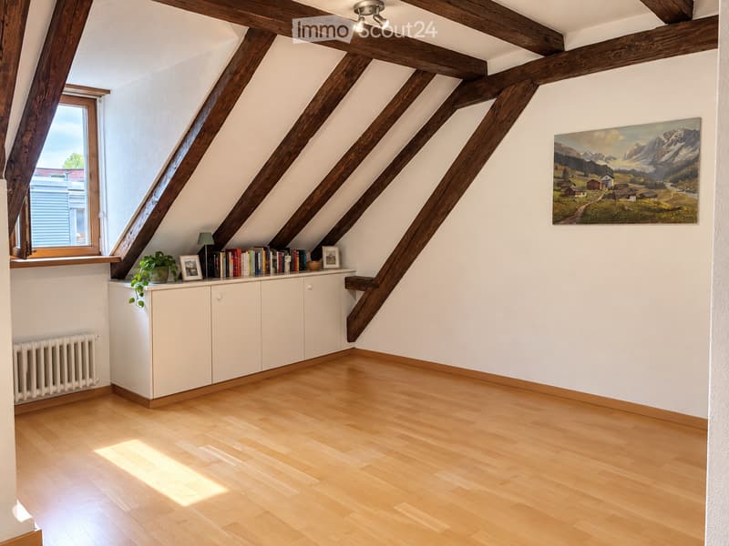 Bright attic room with wooden beams, white walls, wooden floor, large window, cabinet with books and a plant, and a painting on the wall.