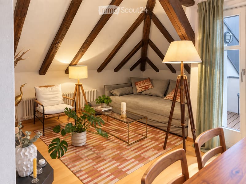Cozy living room with exposed wooden beams, a sofa, armchair, coffee table, floor lamp, and decorative plants.
