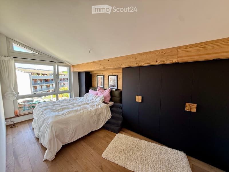 Bright bedroom with wooden floors, large windows, a double bed with white bedding and pink pillows, a dark wardrobe, a white rug, and two picture frames on the wall.