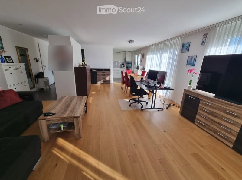 Open-plan living and working space with wooden flooring, large windows, sofa, coffee table, desk, chairs, and TV stand.