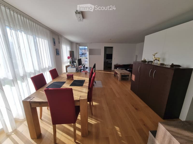 Bright dining area with wooden floor, large window with white curtains, dining table with red chairs, cabinet with decorative items, sofa, coffee table, and TV in the background.