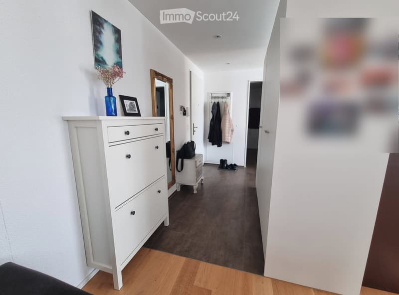 A modern hallway with white walls, wooden flooring, and a white dresser with drawers. A mirror, vase with flowers, and framed picture are on top of the dresser. A coat rack with jackets and a small table with a bag are visible further down the hallway.