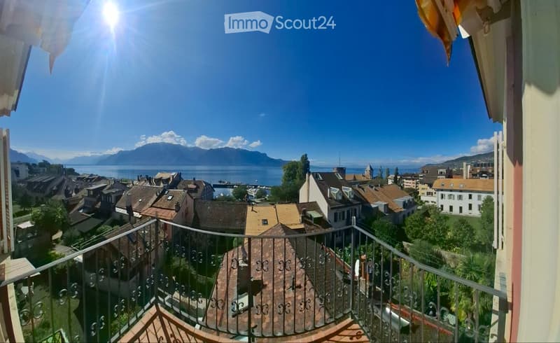 Balcony overlooking a town with a lake and mountains in the background, featuring a metal railing and wooden decking.