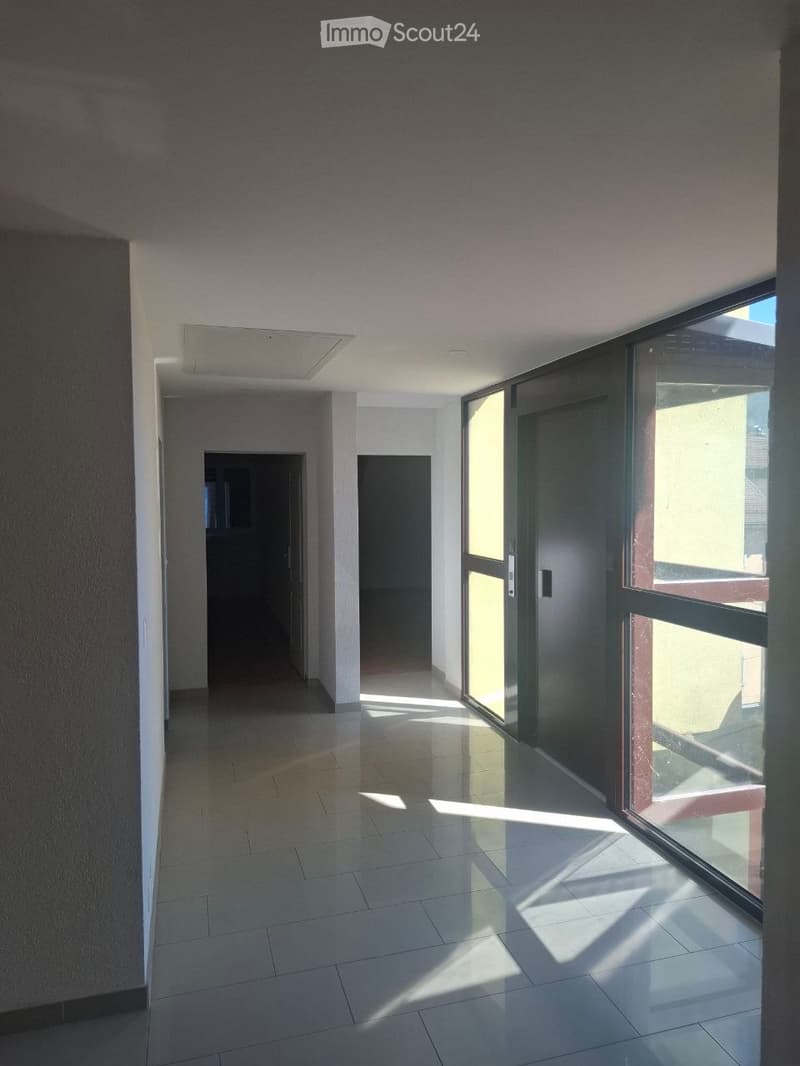 Empty hallway with tiled floor, white walls, and large glass windows allowing natural light to enter. Two doors are visible at the end of the hallway.