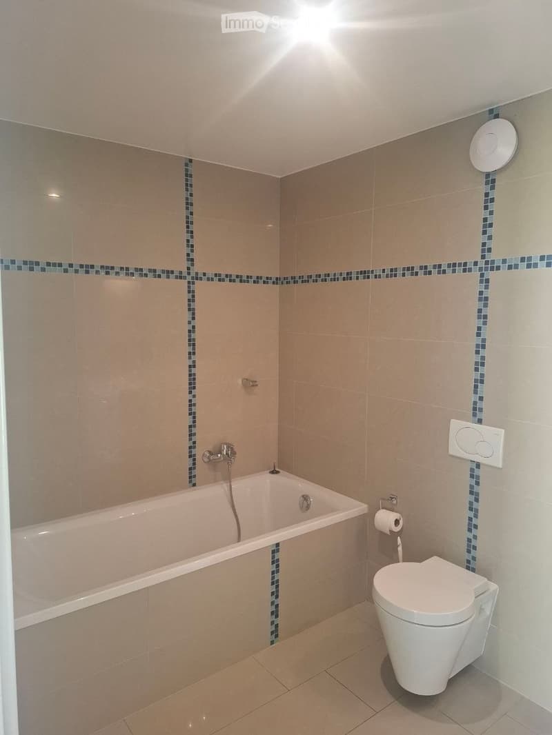 Modern bathroom with beige tiles, a bathtub, toilet, and a faucet. Features a decorative blue mosaic border on the tiles.