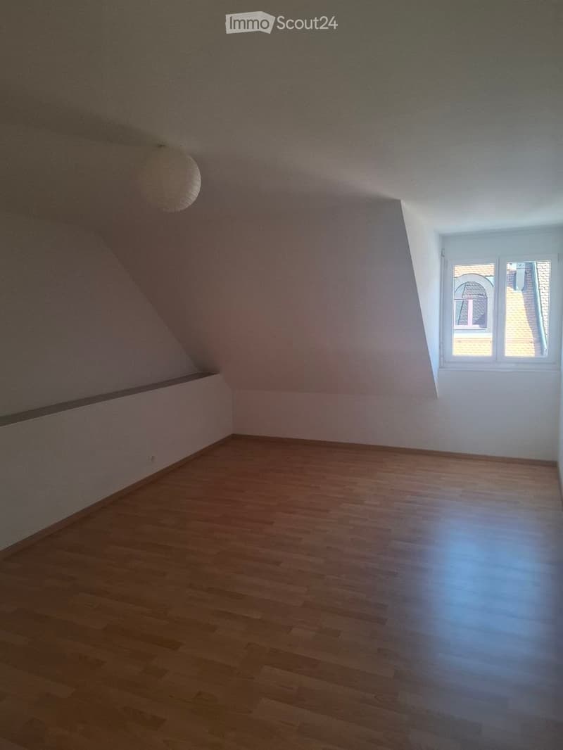 Empty attic room with wooden flooring, slanted ceilings, and a single window letting in natural light.