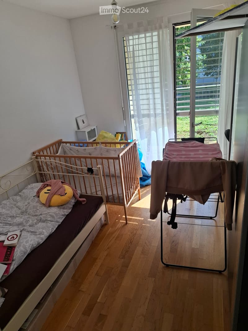 Cozy bedroom with a sofa bed, cushion, mirror, clothing rack, and microwave. The room has wooden flooring and large windows with sheer curtains.