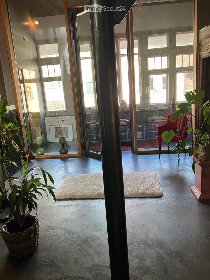 Open-plan living space with dark grey concrete floors, large windows, and glass doors leading to another room. Features include a red chair, a small table with a vase, potted plants, and white rugs.