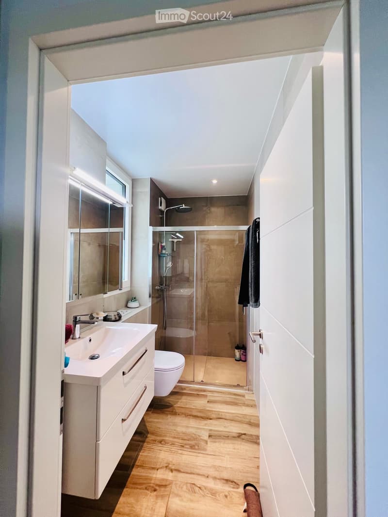 Modern bathroom with a white sink, mirror, toilet, and glass-enclosed shower. Light wooden flooring and white cabinetry.