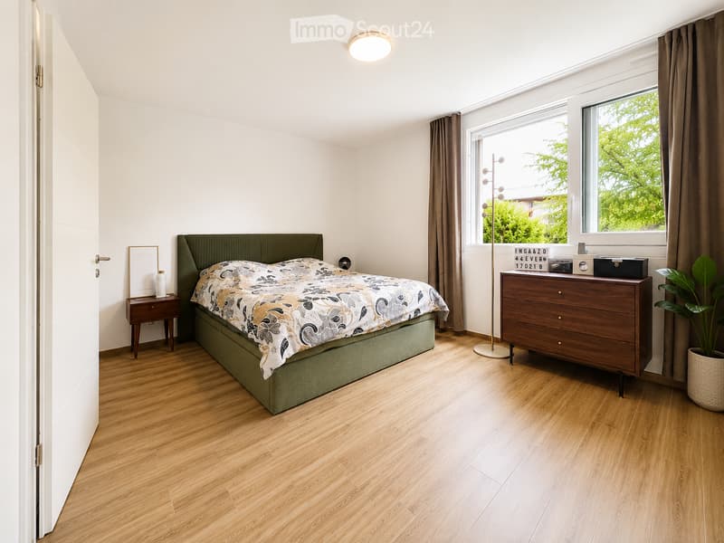 Modern bedroom with wooden flooring, large window with curtains, green upholstered bed, nightstand, dresser, floor lamp, and potted plant.