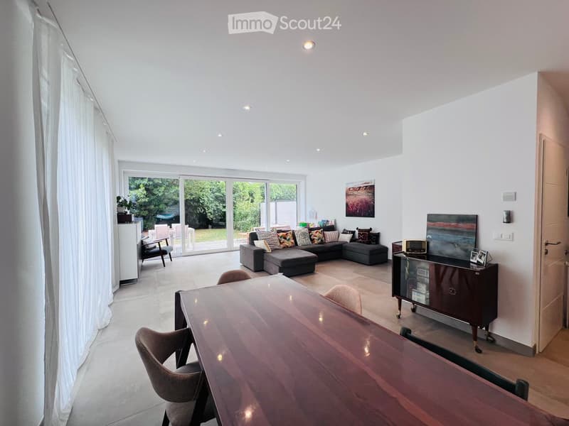 Bright living room with white walls, large windows, and a sliding glass door leading to a patio. Features a grey sectional sofa, coffee table, dining table with chairs, and a cabinet with a TV on top.