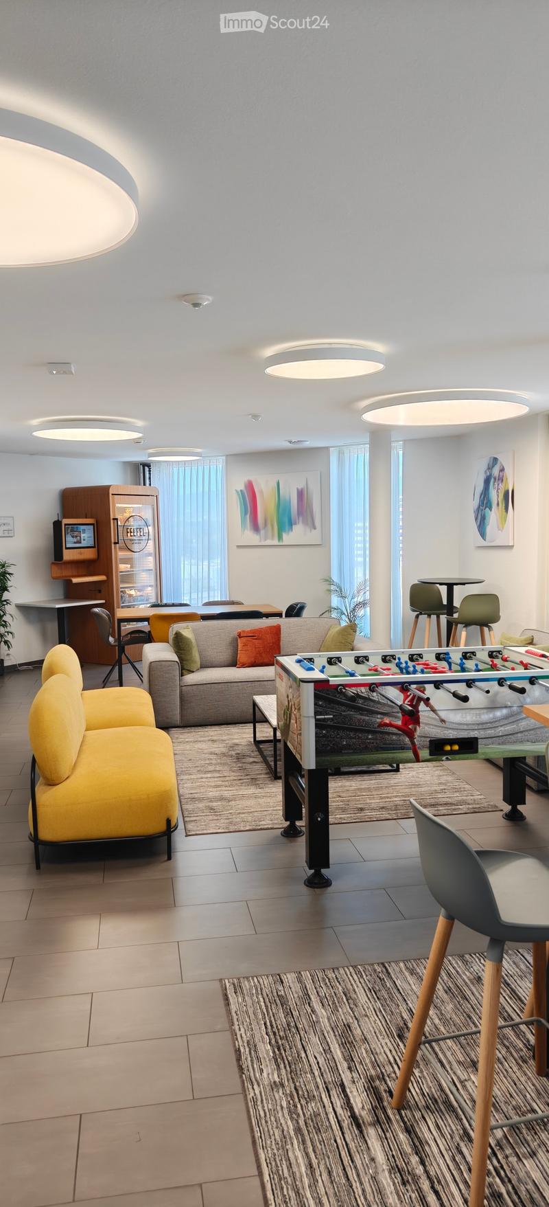 Modern living room with a sofa, football table, chairs, and a screen. The room features large windows with curtains, modern lighting, and colorful artwork on the walls.