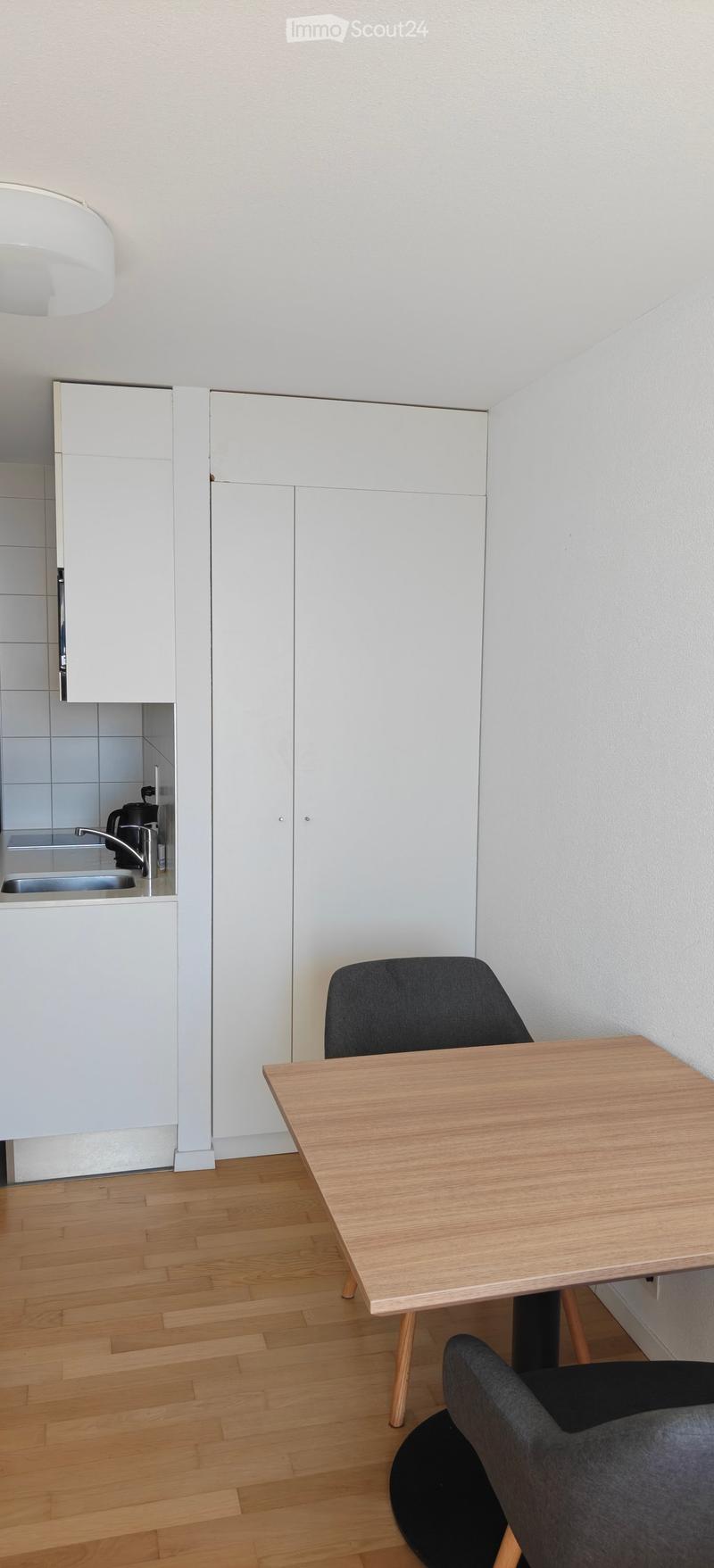 A modern, minimalist kitchen area with white cabinets, a wooden dining table, and a grey chair. The floor is made of light wooden planks, and there is a small sink area visible in the background.