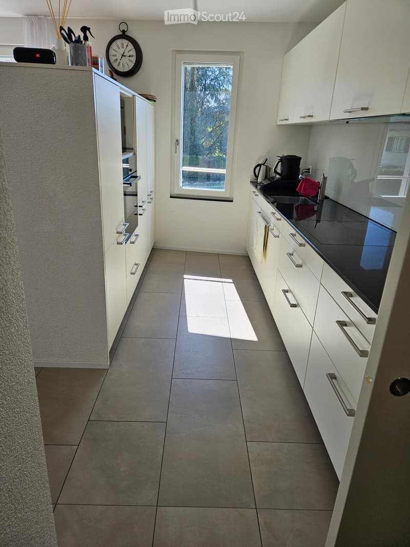 Modern kitchen with white cabinets, stainless steel handles, and a black countertop. Features a window, a clock, and various kitchen items on the counter.