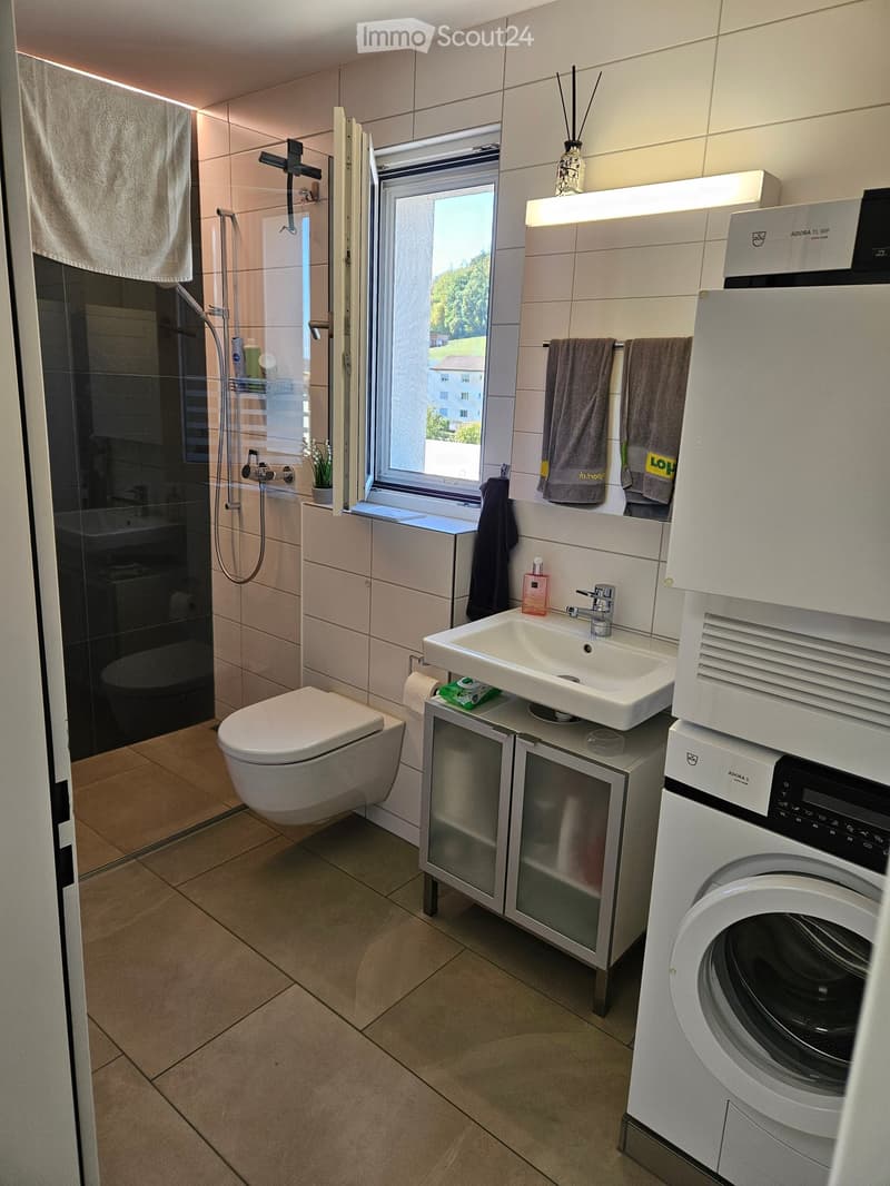 Modern bathroom with a toilet, sink, shower, and washing machine. Features include a large window with a view, a mirror, and a towel rack with towels.