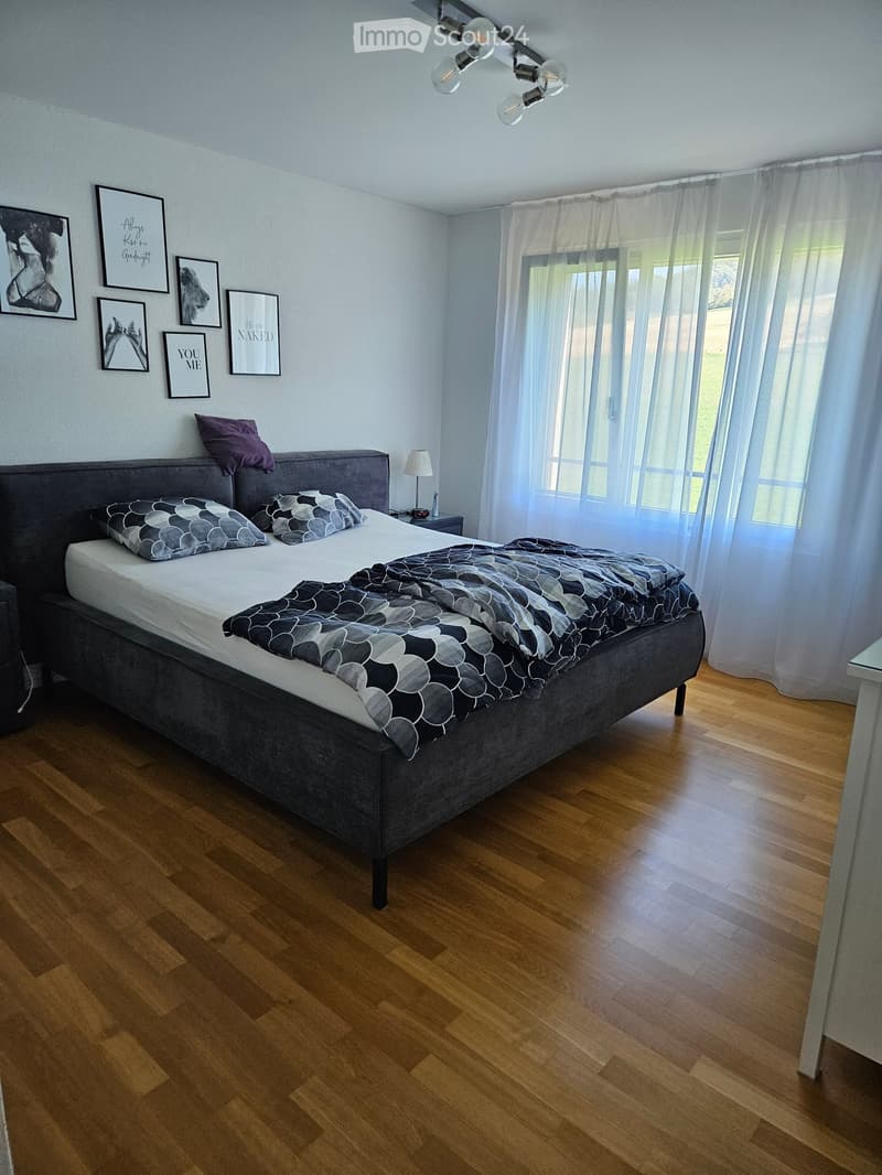 Modern bedroom with a large bed, grey headboard, patterned bedding, nightstand with lamp, and several picture frames on the wall. Wooden flooring and large window with sheer curtains.