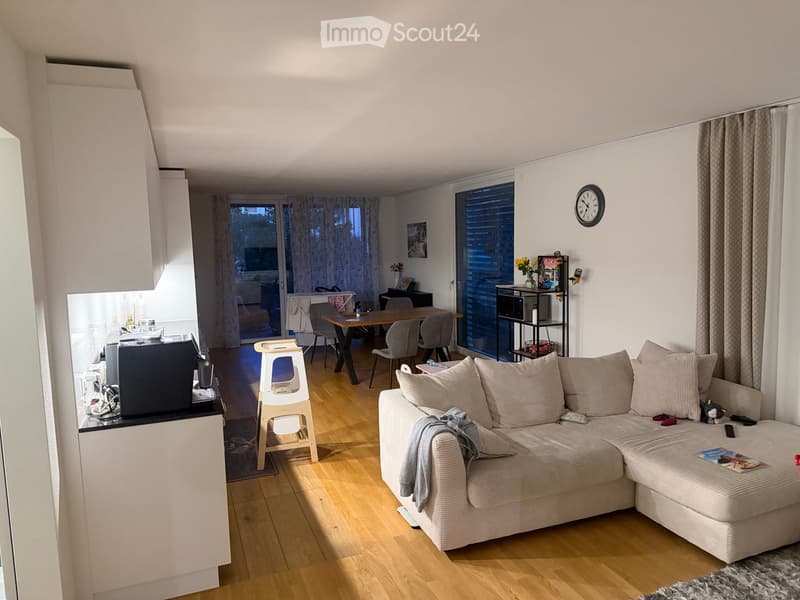 A modern living room with a sectional sofa, a small wooden stool, a dining table with chairs, a television, and a clock on the wall. The room features wooden flooring and large windows with blinds.
