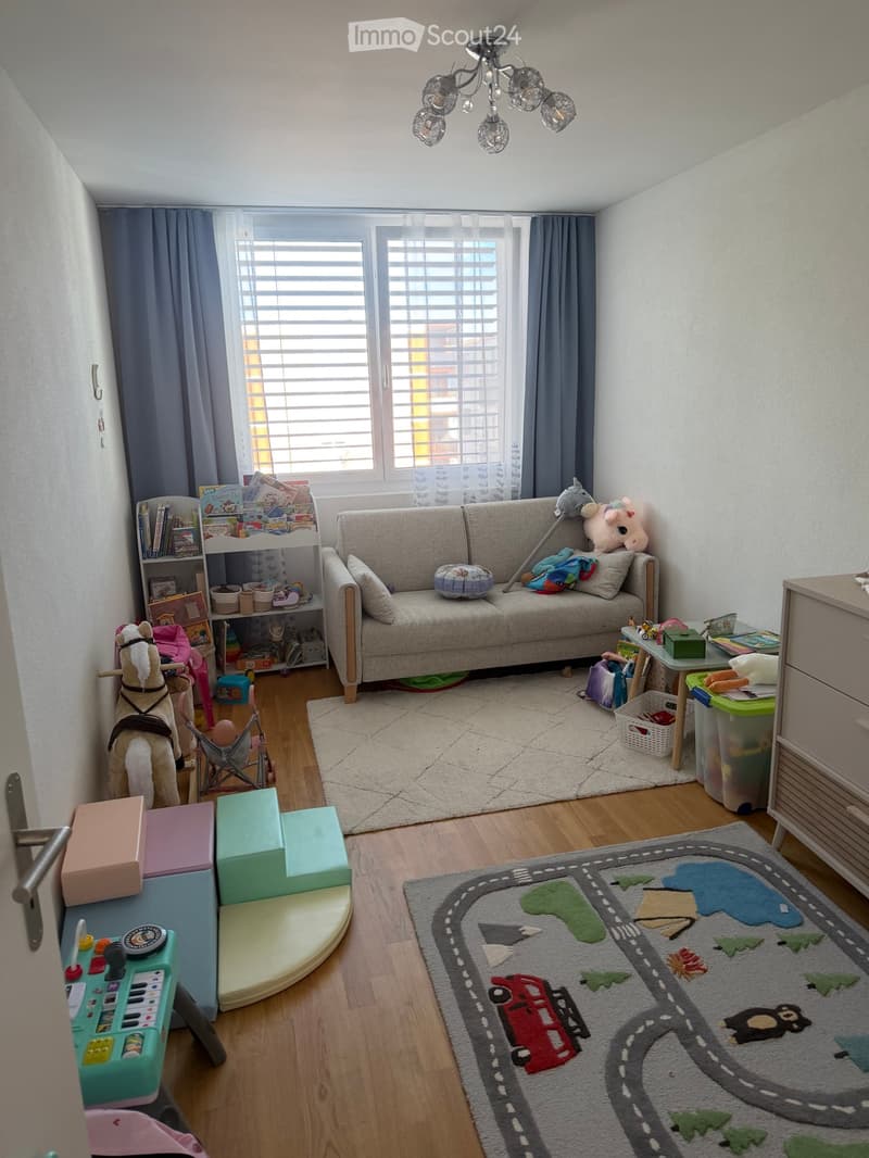 A well-lit children's room with a grey sofa, bookshelf, toy horse, play table, play mat, and various toys.