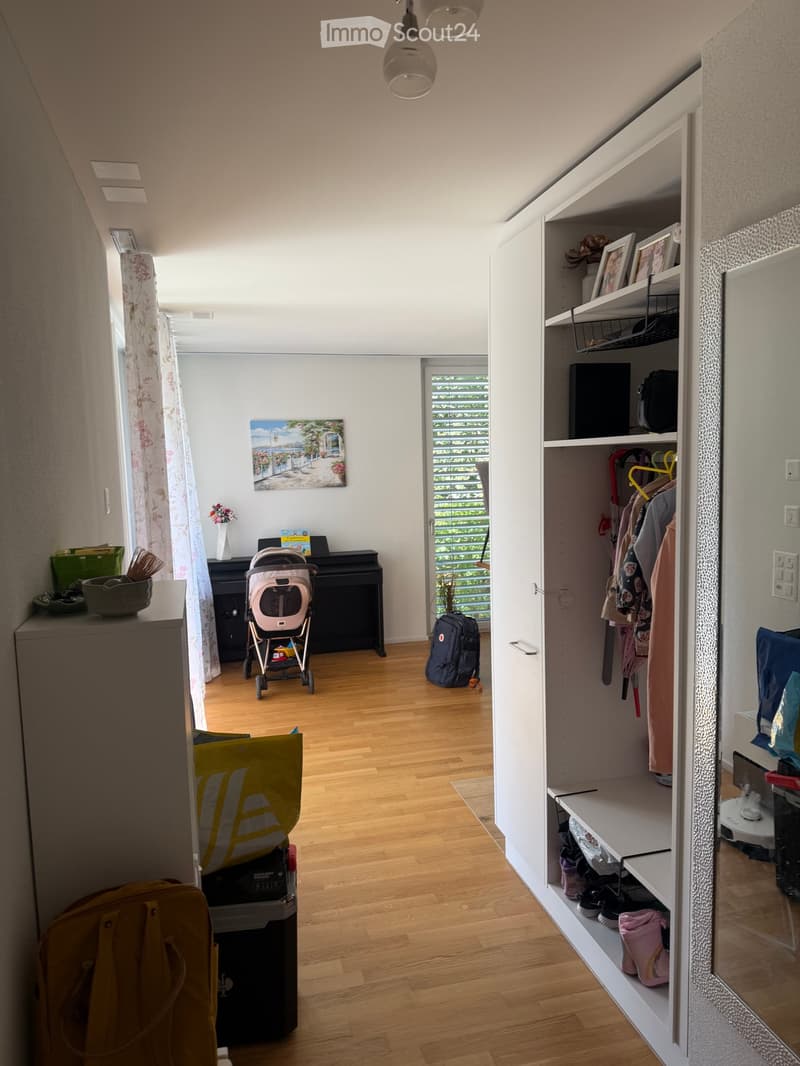 A well-lit hallway with wooden flooring, a piano, a baby stroller, and an open closet with clothes and shoes.