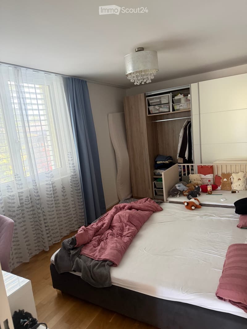 A well-lit bedroom with a large bed, a wardrobe, and a window with curtains. The bed is partially made with blankets and pillows, and there are toys on a changing table nearby.