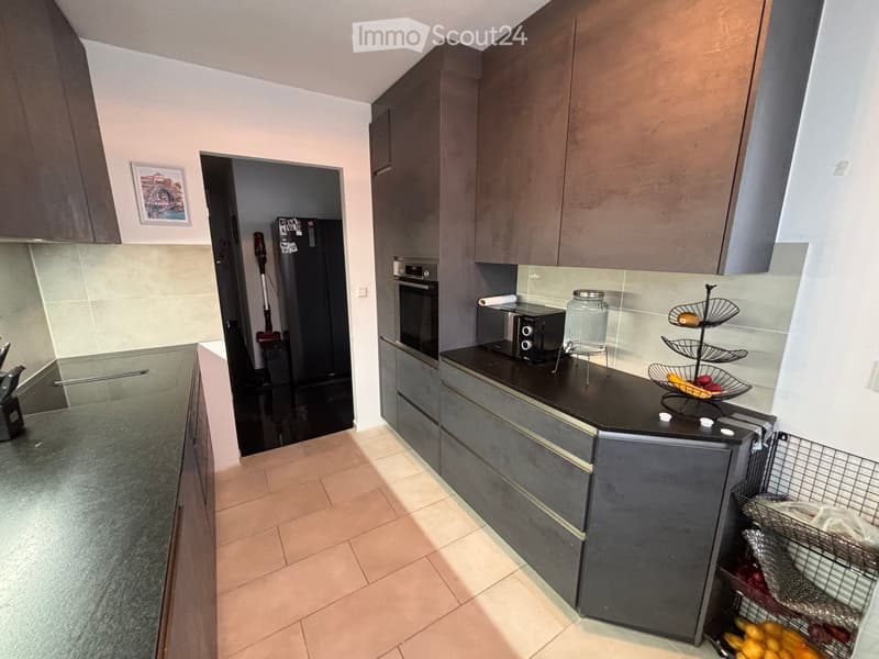 Modern kitchen with grey cabinets, black countertops, and stainless steel appliances. Features a refrigerator, oven, microwave, and a fruit basket on the counter.