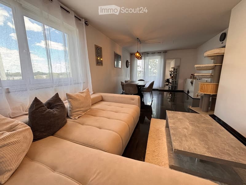 Modern living room with a white sectional sofa, coffee table, dining area with chairs, and a small kitchen area visible in the background.
