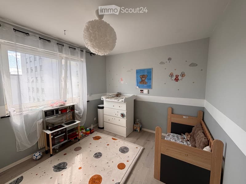 A well-lit children's bedroom with a bunk bed, dresser, small table, and various toys. The room features a space-themed rug and playful wall decorations.