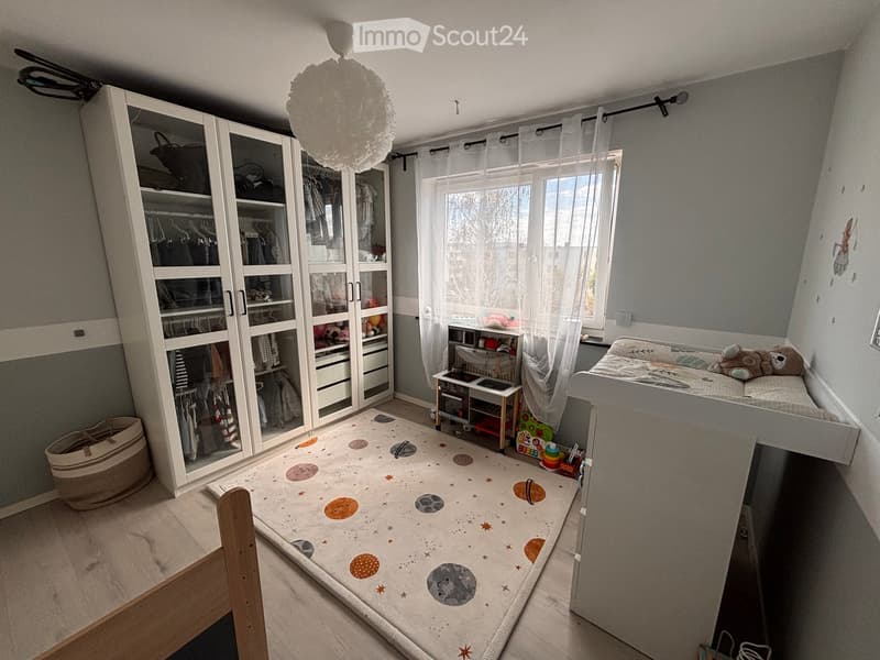 A well-lit bedroom with a large wardrobe, a loft bed, a small table with toys, and a colorful rug on the floor.